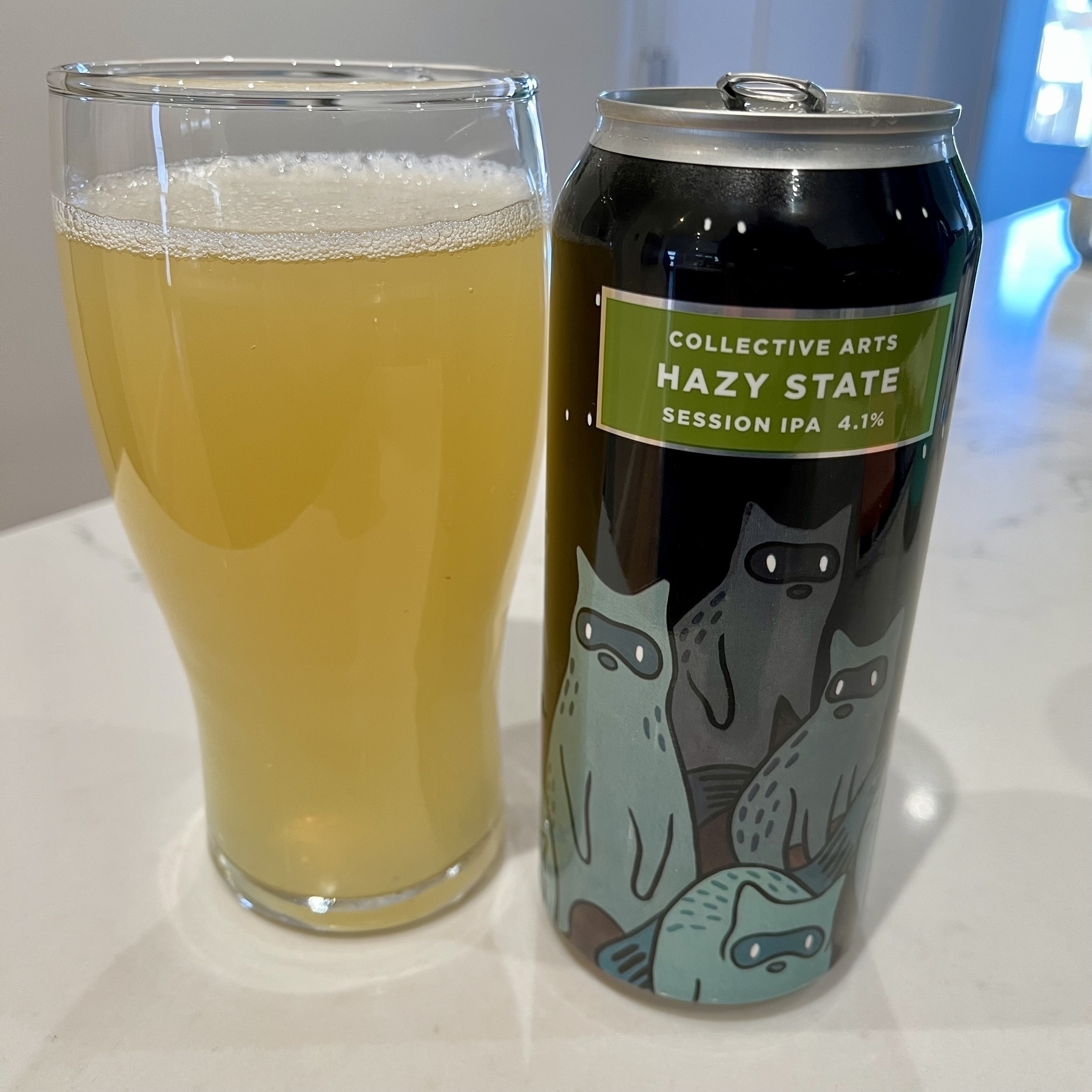 A hazy yellow beverage is poured into a glass next to a can labeled "COLLECTIVE ARTS HAZY STATE SESSION IPA 4.1%" with stylized cat illustrations on a kitchen counter.